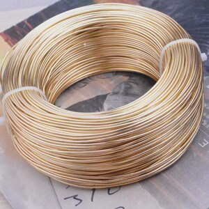 Pode incluir: Uma bobina de fio dourado, enrolada firmemente e presa com laços brancos. O fio parece liso e brilhante, adequado para artesanato ou joalheria. O fundo é uma superfície impressa com texto.
