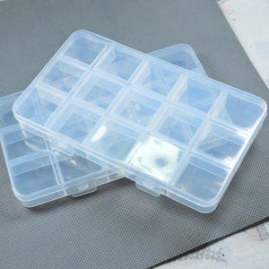 Puede incluir: Un contenedor de almacenamiento rectangular de plástico transparente con quince compartimentos individuales dispuestos en cuadrícula. El organizador cuenta con una tapa abatible con un mecanismo de cierre seguro y vista clara.