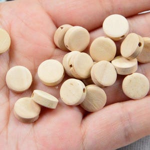 以下が含まれることがあります： 自然な淡いタン色の、小さくて丸い未完成の木製ビーズのコレクション。各ビーズには、ひもを通すための穴が中央に開いています。ビーズは手に散らばっており、そのサイズと形状を示しています。