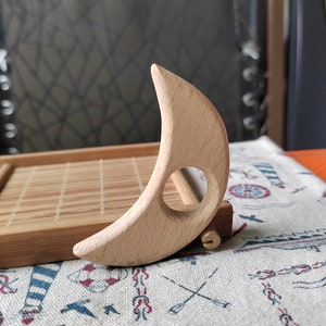 May include: A wooden crescent moon shaped tool with a hole in the center. The tool is made of light brown wood and is sitting on a fabric surface with a nautical pattern.