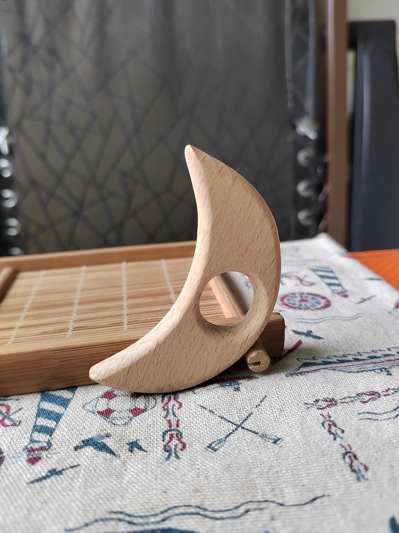 May include: A wooden crescent moon shaped tool with a hole in the center. The tool is made of light brown wood and is sitting on a fabric surface with a nautical pattern.