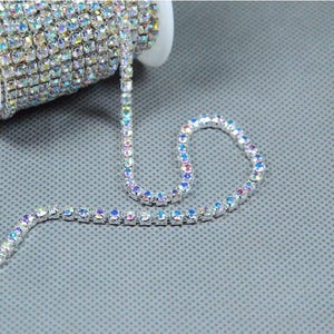 Puede incluir: Un rollo de ribete de pedrería iridiscente con un hilo desenrollado. Los diamantes de imitación son transparentes con un brillo de arcoíris, engastados en un metal plateado. El ribete está sobre una bobina blanca sobre un fondo gris.