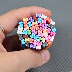 Puede incluir: Un conjunto de cañas de arcilla polimérica de colores, cada una con un diseño de lazo. Los lazos son de varios colores, incluyendo rosa, azul, verde y blanco. Las cañas se sostienen en una mano sobre un fondo gris.