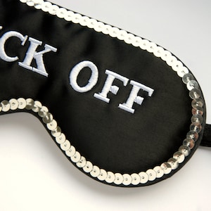 以下が含まれることがあります： 縁に銀色のスパンコールが付いた黒いアイマスク。マスクには「F**k Off」という文字が白い糸で刺繍されている。