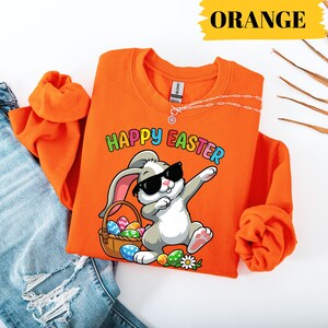 Könnte beinhalten: Leuchtend orangefarbenes Sweatshirt mit dem bunten Schriftzug "HAPPY EASTER". Die Grafik zeigt ein graues Häschen mit Sonnenbrille, das dabbt, neben einem Korb mit Ostereiern. Eine silberne Halskette ist sichtbar.