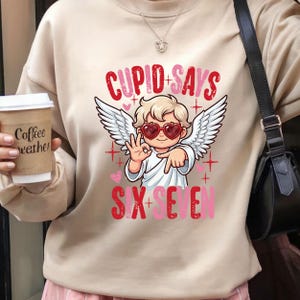 May include: Beige sweatshirt with a graphic of Cupid wearing heart-shaped sunglasses and angel wings. The text "Cupid Says" and "Six Seven" are in red and pink. A coffee cup with "Coffee weather" is held in the hand.