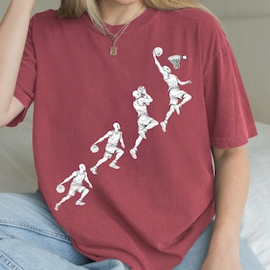 Puede incluir: Camiseta de color burdeos con un gráfico blanco que representa la secuencia de un mate de un jugador de baloncesto. El diseño muestra al jugador botando, saltando y lanzando el balón hacia el aro. La camiseta es de material suave.
