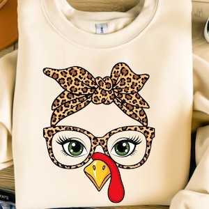 May include: A cream-colored sweatshirt featuring a cartoon turkey face. The turkey has a leopard print bow, glasses, and green eyes. The beak is yellow, and the wattle is red. A fun, festive design for Thanksgiving.