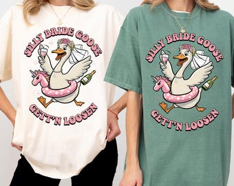 May include: Two t-shirts, one cream and one sage green, featuring a cartoon goose wearing a bridal veil and a pink unicorn floatie. The text reads "Silly Bride Goose, Gett'n Loosen". The goose holds a wine glass and a bottle.