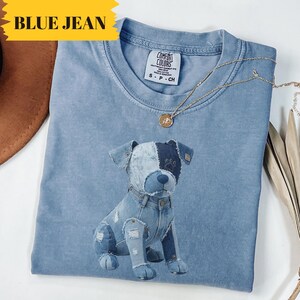 May include: A light blue Comfort Colors t-shirt featuring a denim dog graphic. The dog has patches of light and dark blue denim. A gold necklace with a pendant rests on the shirt. The words "BLUE JEAN" are in a yellow banner.