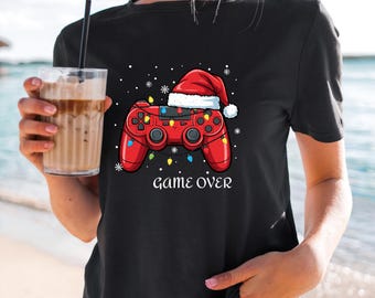 Könnte beinhalten: Schwarzes T-Shirt mit einem weihnachtlichen Design. Es zeigt einen roten Gamecontroller, der mit einem Weihnachtsmannhut und Lichterketten geschmückt ist. Die Worte "GAME OVER" sind darunter gedruckt. Schneeflocken vervollständigen das Design.