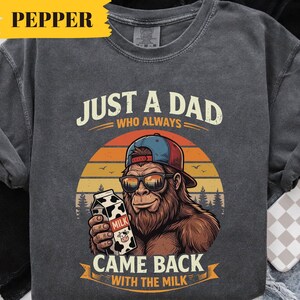 May include: Dark gray sweatshirt with a graphic of a Bigfoot wearing sunglasses and a baseball cap, holding a milk carton. Text reads "JUST A DAD WHO ALWAYS CAME BACK WITH THE MILK". The design features a sunset and trees.