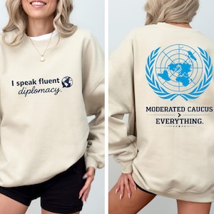 Puede incluir: Sudadera beige con el texto "I speak fluent diplomacy" y un gráfico de globo terráqueo en la parte delantera. La parte trasera presenta el emblema azul de las Naciones Unidas y las palabras "Moderated Caucus > Everything."