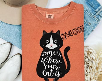 Può includere: T-shirt arancione Comfort Colors con un'illustrazione di gatto in bianco e nero e la scritta "Home is Where Your Cat is". La parola "Meow" è scritta in un font stilizzato. La maglietta è abbinata a jeans blu e stivali bianchi.