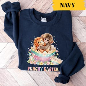 Könnte beinhalten: Marineblaues Sweatshirt mit einem farbenfrohen Aufdruck von zwei Affen, die sich in einem zerbrochenen Osterei umarmen. Das Design umfasst Pastellfarben, Blumen und den Text "PUNCHY EASTER". Das Wort "NAVY" ist in Gelb gedruckt.