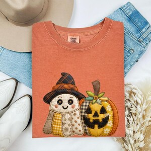 May include: Orange Comfort Colors t-shirt featuring a patchwork ghost wearing a witch hat and a jack-o'-lantern. The ghost has button eyes and a smiling mouth. The pumpkin has a smiling face and a green stem. The shirt is folded on a white surface.