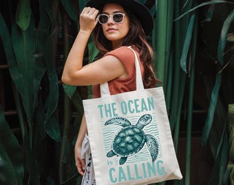 May include: Cream-colored tote bag with the text "THE OCEAN IS CALLING" in light blue. The bag features a teal sea turtle illustration. The bag has white handles and is being held in front of a lush green background.