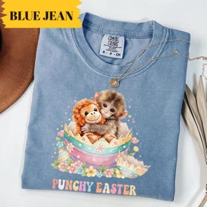 Könnte beinhalten: Hellblaues Comfort Colors T-Shirt mit einer Grafik von zwei Affen, die sich in einem dekorierten Osterei umarmen. Der Text "PUNCHY EASTER" steht darunter. Eine goldene Halskette liegt auf dem Shirt. Die Worte "BLUE JEAN" stehen in einem gelben Banner.