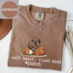 May include: Espresso-colored Comfort Colors t-shirt with a graphic of a small, brown monkey resting on a larger, orange-brown monkey. The text "soft heart, tired mind #punch" is printed below the graphic. A silver necklace with a small pendant is draped on the shirt.