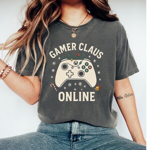 May include: Dark gray t-shirt with the words "Gamer Claus Online" and a cream-colored controller graphic. The controller has Christmas-themed accents. The shirt is a casual, festive garment.