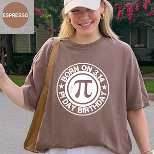 Puede incluir: Camiseta marrón con un gráfico blanco que dice "BORN ON 3.14 PI DAY BIRTHDAY" alrededor del símbolo Pi. La camiseta es de manga corta y cuello redondo. La imagen también muestra una gorra beige y un bolso de hombro marrón.