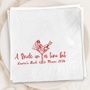 May include: White cocktail napkins with a red illustration of a bride in a martini glass. The text reads "A Bride in a tini bit" with "Laura's Bach Miami 2026" below the illustration. The napkins are square and have a subtle diamond pattern.