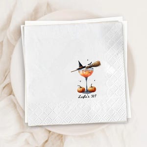 May include: A white square paper napkin featuring a printed illustration of an orange cocktail in a stemmed glass, topped with a witch hat and broomstick. Two small orange pumpkins sit at the base, with the text Layla's 30! printed below.