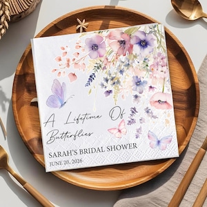 May include: White square paper napkin with floral and butterfly design. The napkin features a watercolor illustration of purple, pink, and blue flowers and butterflies. Text reads "A Lifetime Of Butterflies" and "SARAH'S BRIDAL SHOWER JUNE 20, 2026".