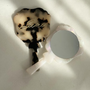 May include: Two hand mirrors. One has a tortoiseshell pattern with the name "Simi" in gold script. The other mirror is white with a pearlescent sheen and a round mirror surface. Both mirrors have a handle.