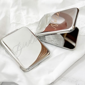 Puede incluir: Espejos compactos plateados con nombres grabados. Un espejo está abierto, revelando un interior marrón. La palabra "Bride" está grabada en un espejo, y "Nola" en el otro. Los espejos están sobre una superficie blanca.