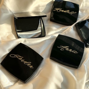 Op de afbeelding: Zwarte compacte spiegels met gouden letters die namen spellen, waaronder "Kendall", "Annie" en "Jordan". Eén spiegel is open en onthult een reflecterend oppervlak. De spiegels zijn gerangschikt op een witte, zijdeachtige stof.