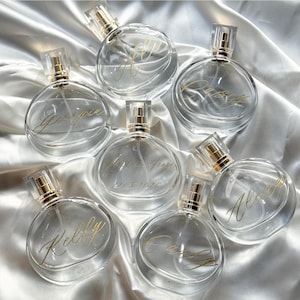 Peut inclure: Plusieurs flacons de parfum en verre transparent avec des vaporisateurs dorés et des inscriptions dorées personnalisées. Les bouteilles sont disposées sur une surface en satin blanc. Certaines bouteilles portent des noms tels que "Kelly" et "Georgia". Une bouteille porte l'inscription "Mark & Hannah 12.03.2020".