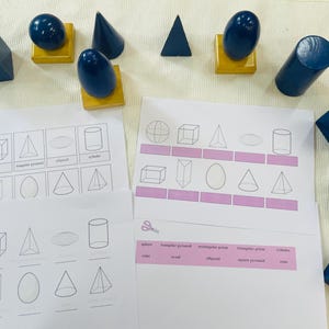 Könnte beinhalten: Lehrset mit dunkelblauen geometrischen Formen, darunter Würfel, Kegel und Zylinder, sowie Arbeitsblätter. Die Formen sind auf einer hellen Oberfläche angeordnet, ideal zum Lernen.