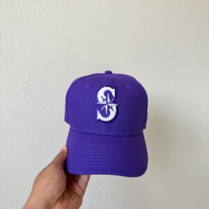 May include: A purple baseball cap with a white and blue embroidered logo. The cap features a curved brim and a structured crown. The logo is a stylized letter "S" with a compass rose design.