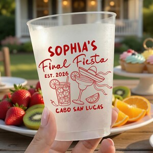 May include: A clear plastic cup with red text that reads "Sophia's Final Fiesta" and "Est. 2026". The cup features illustrations of a margarita, a cocktail, and a sombrero. The cup is held in front of a table with fruit and cupcakes.