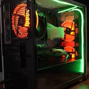 May include: A black computer case with a clear side panel, revealing the interior components. The case has green LED lights around the edge and orange LED fans inside. The case is sitting on a wooden surface.