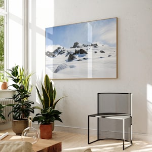 May include: A framed landscape photograph of a snow-covered mountain range with dark rock formations. The photograph is displayed on a white wall in a room with plants and a modern black chair.