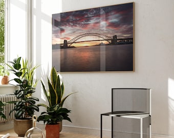 May include: A framed print of a bridge at sunset, with a vibrant sky of red and orange hues. The bridge spans across a body of water, reflecting the warm colors. The print is displayed on a white wall, with a modern black chair and potted plants in the foreground.