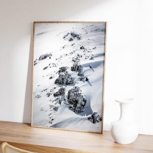 May include: A framed print of a snow-covered mountain slope with dark rocks. The artwork is displayed on a wooden surface, next to a white vase. The frame is a light wood color.