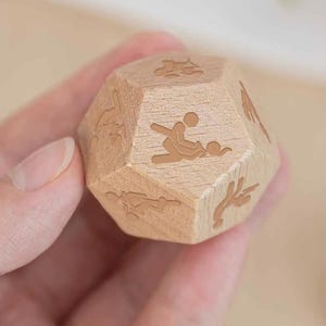 May include: A wooden, multi-sided die with etched illustrations. The die is held in a hand and features various figures and symbols on its faces. The die is made of light-coloured wood.
