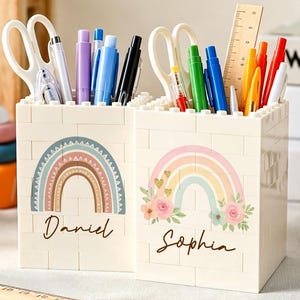 May include: Two white rectangular pencil holders constructed from interlocking plastic bricks. Each holder features a printed rainbow design and a name, Daniel and Sophia, in black script. They are filled with various pens, scissors, and a ruler.