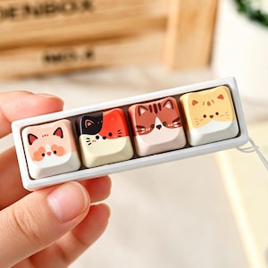 May include: A white rectangular mechanical keyboard switch tester base holding four individual keycaps. Each keycap features a distinct cartoon cat face design in shades of orange, black, white, and brown, with a white lanyard attached to the side.