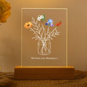 May include: A rectangular acrylic night light with a wooden base featuring a white line drawing of a mason jar filled with flowers. The flowers are colored orange, white, blue, and red, with names written on the stems. The text reads We love you Mummy x.