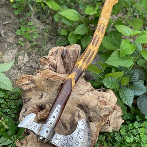Puede incluir: Un hacha decorativa con una hoja plateada y un mango de madera. La hoja presenta intrincados diseños grabados y el mango es una combinación de madera oscura y clara con detalles dorados. El hacha se exhibe al aire libre sobre un fondo de follaje verde.