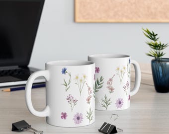 May include: Two white ceramic mugs with a floral design. The mugs feature various watercolor flowers in shades of purple, pink, and blue, along with green leaves. The mugs are on a light-colored surface, with a laptop and office supplies in the background.