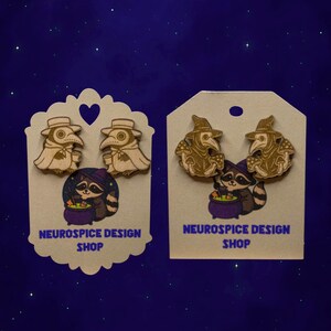 May include: Wooden earrings featuring a plague doctor design with a raccoon and a cauldron. The earrings are on a decorative card with the text "NEUROSPICE DESIGN SHOP". The earrings are light brown and the raccoon is wearing a purple hat.