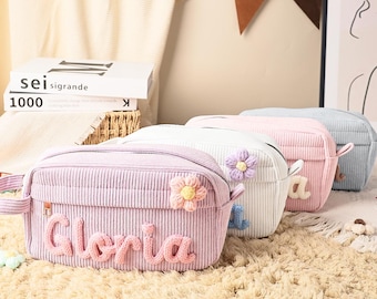 May include: A row of four rectangular corduroy pencil cases in lavender, white, pink, and light blue. Each case features a side handle, a zipper closure, and a decorative crocheted flower. The lavender case has the name Gloria in raised letters.