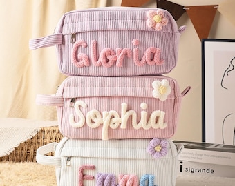 May include: Three rectangular corduroy pouches stacked vertically. The top pouch is lavender with the name Gloria in pink yarn. The middle pouch is light pink with the name Sophia in white yarn. The bottom pouch is white with the name Emma in multicolored yarn. Each pouch features a small crocheted flower accent.