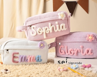 May include: Three rectangular corduroy pouches in light pink, white, and lavender. Each pouch features a zippered closure, a small crocheted flower accent, and a personalized name in raised, soft-textured lettering reading Sophia, Emma, or Gloria.