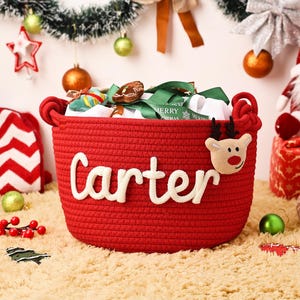 May include: A red woven basket with the name "Carter" in white script. The basket is decorated with a reindeer and filled with gifts and ribbons. Christmas ornaments and decorations surround the basket.